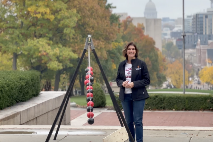 a woman stands in front of an apparatus that looks like a swingset but has 15 bocce balls in a line, alternating red and black, one behind the other. The balls are on strings of different lengths, too, so they aren't in a straight line, they gradually get shorter the further back they are in the apparatus